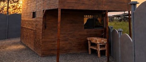 A two-story wooden cabin with a sloped roof, a stone wall, and a gravel ground.