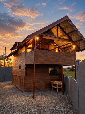 A two-story wooden cabin with a sloped roof, a stone wall, and a gravel ground. - Shanti Cabin | Hot Tub and Romantic Getaway (Venecia)