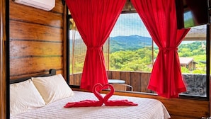 Habitació d'hotel amb un llit, un ventilador de sostre, cortines vermelles, una televisió i vistes a vegetació.