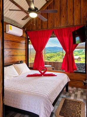 Ein Hotelzimmer mit Bett, Deckenventilator, roten Vorhängen, Fernseher und Blick ins Grüne.