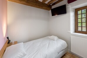 Ein Einzelbett mit weißer Bettwäsche in einem Zimmer mit weißen Wänden, einer Holzbalkendecke und einem Fenster mit Blick auf Bäume.