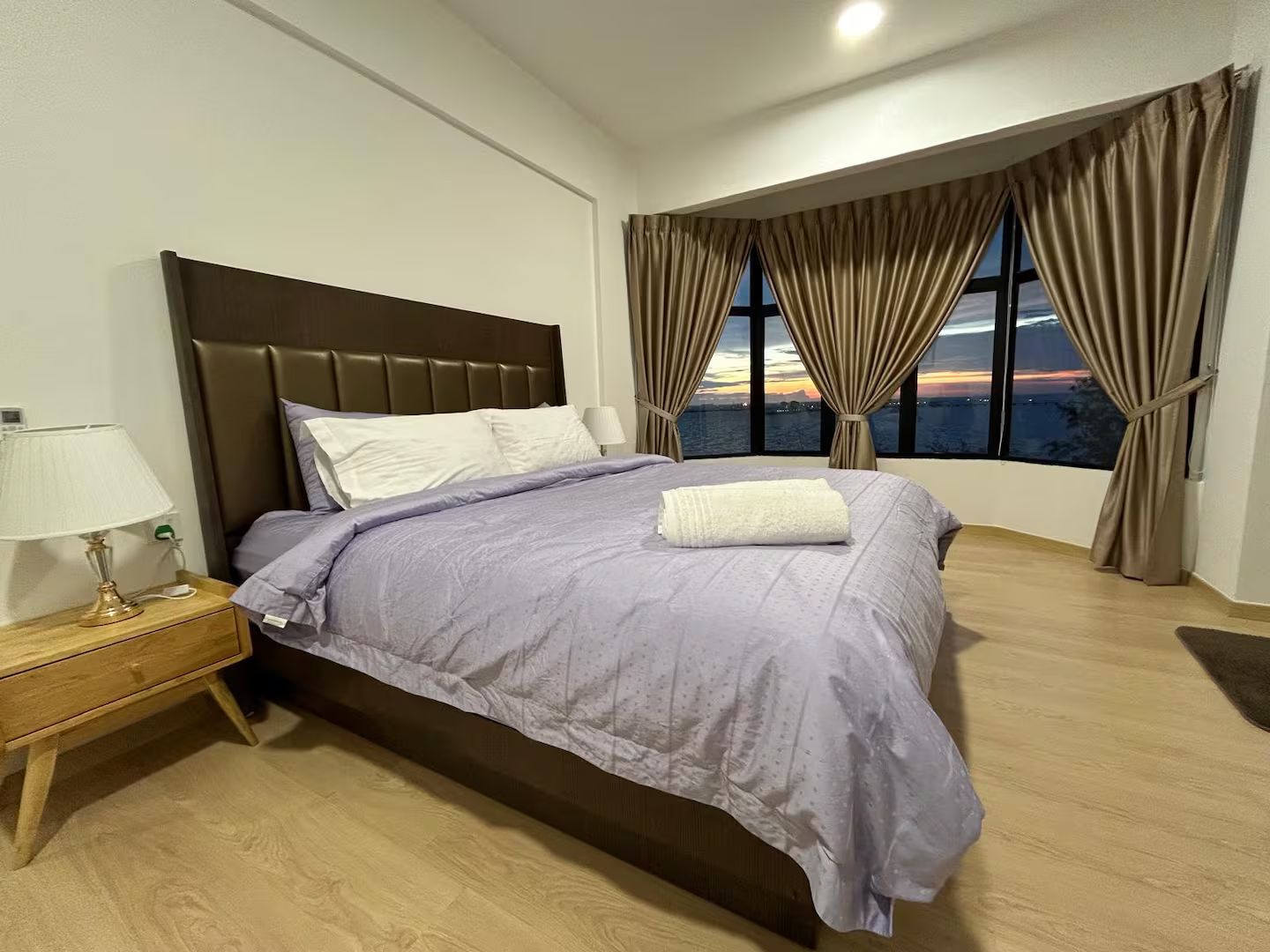 Ein Schlafzimmer mit Bett, Nachttisch, Lampe und Blick auf die Stadt durch große Fenster.