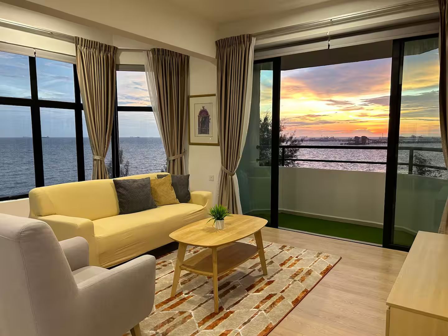 Ein modernes Wohnzimmer mit Sofa, Sessel und Couchtisch, großen Fenstern mit Vorhängen und Blick auf den Sonnenuntergang über dem Wasser.