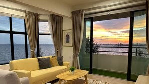 A modern living room with a sofa, armchair, and coffee table, large windows with curtains, and a view of the sunset over the water.