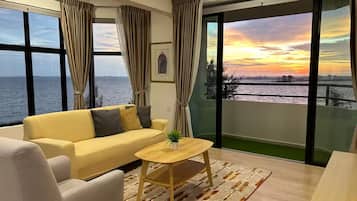 Ein modernes Wohnzimmer mit Sofa, Sessel und Couchtisch, großen Fenstern mit Vorhängen und Blick auf den Sonnenuntergang über dem Wasser.