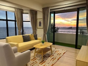Una sala moderna con sofá, sillón y mesa de centro, ventanas amplias con cortinas, y vista al atardecer sobre el agua.