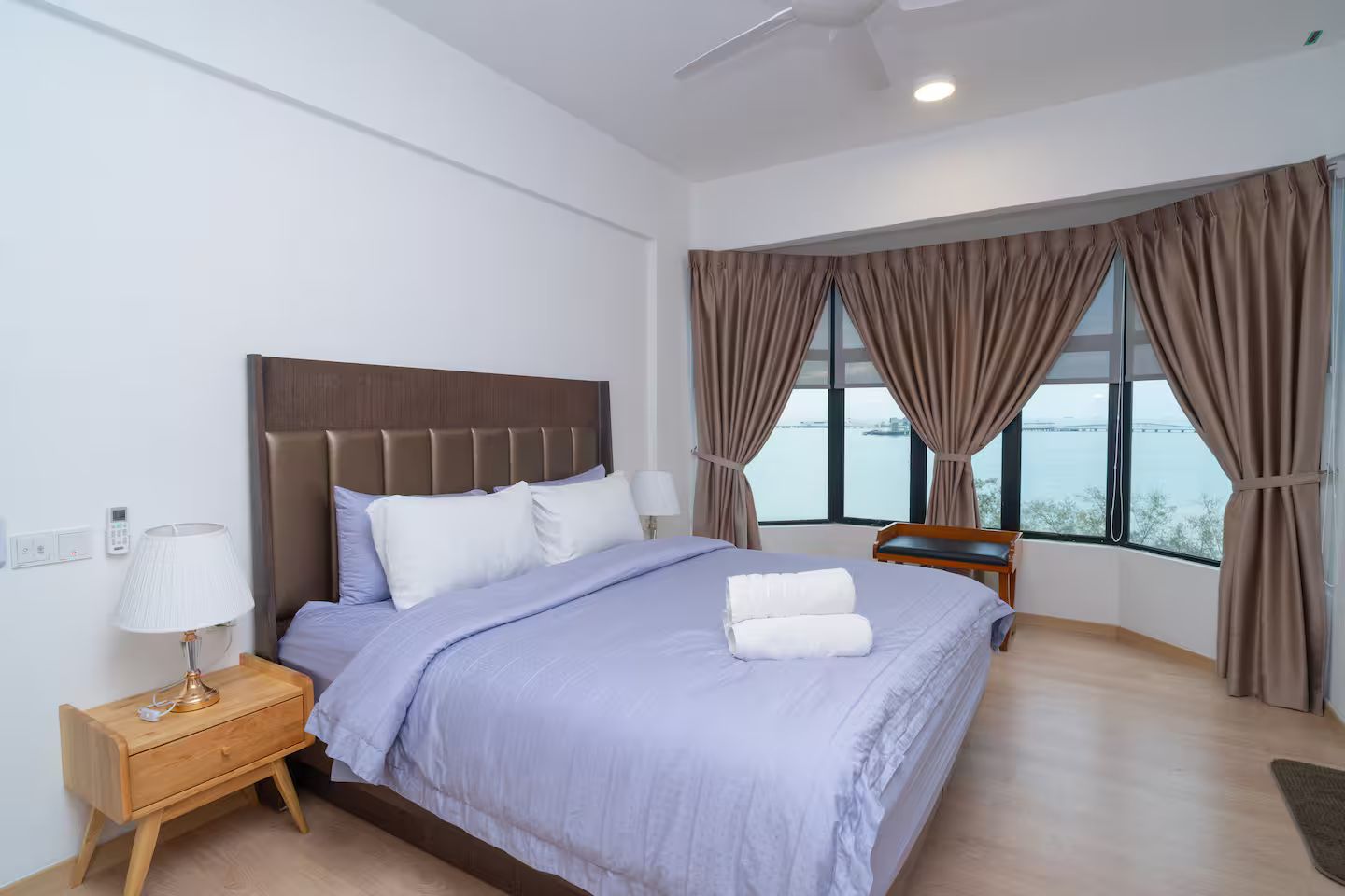 Habitación de hotel con cama, mesita de noche, lámpara y vista al mar a través de amplias ventanas.