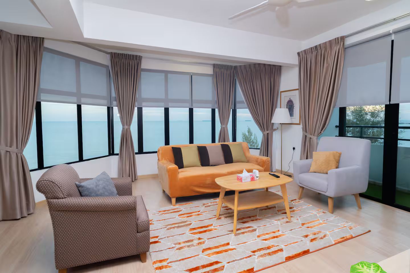 Una sala de estar moderna con sofá, sillón y mesa de centro, ventanas amplias con cortinas y vista al mar.