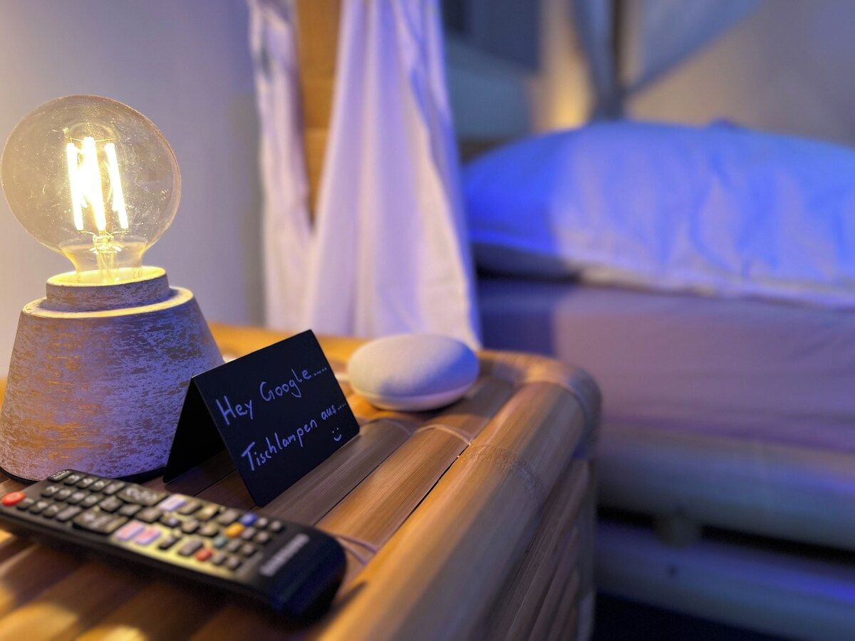 Ein Bett mit weißer Bettdecke, ein Nachttisch mit Lampe, einer Fernbedienung und einem Schild mit handgeschriebenem Text.