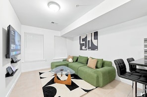 A modern living room with a green sofa, a glass coffee table, a flat-screen TV, and a dining area with a table and chairs. - A Cozy Boho Oasis | 1BR Center City Philly (Philadelphia)