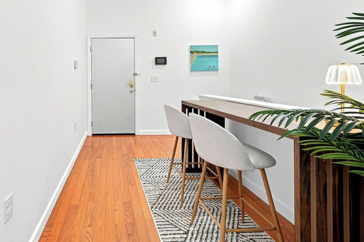 Un coin repas avec une table en bois, deux chaises, un tapis à motifs, une porte, un interrupteur fixé au mur et une plante en pot.