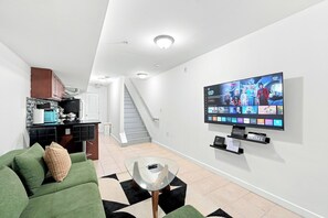 A modern living room with a flat-screen TV mounted on the wall, a green sofa, a glass coffee table, and a kitchen area with cabinets and a countertop. - A Cozy Boho Oasis | 1BR Center City Philly (Philadelphia)