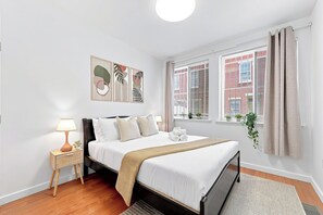 A bedroom with a bed, bedside table, lamp, artwork, window with curtains, and a view of buildings. - A Cozy Boho Oasis | 1BR Center City Philly (Philadelphia)
