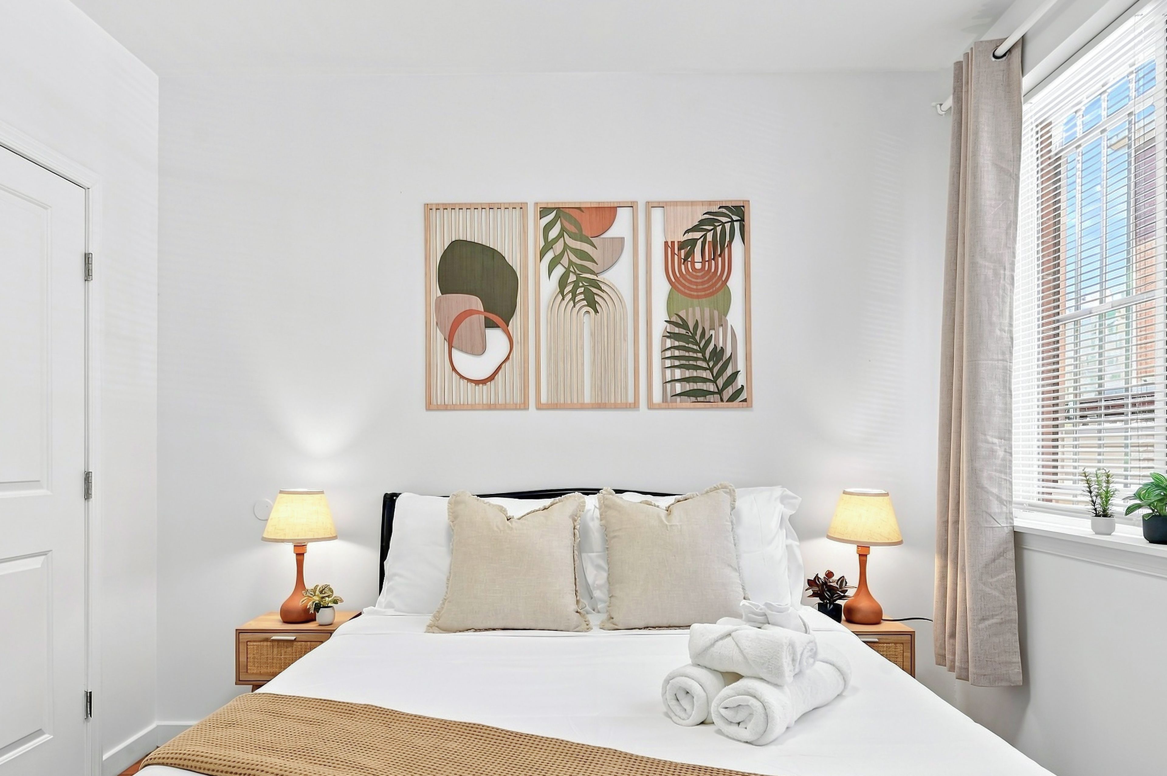 Habitación de hotel con cama, dos lámparas de noche, ventana con persianas y una pared con tres cuadros enmarcados.