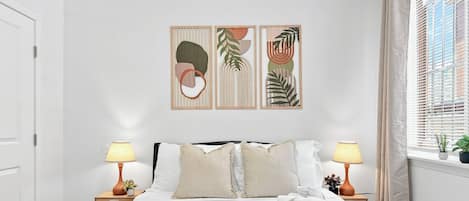 Habitación de hotel con cama, dos lámparas de noche, ventana con persianas y una pared con tres cuadros enmarcados.