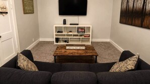 Sala de estar con un televisor de pantalla plana montado en la pared, un mueble blanco debajo, una mesa de centro en el centro y un sofá de color oscuro con cojines estampados.