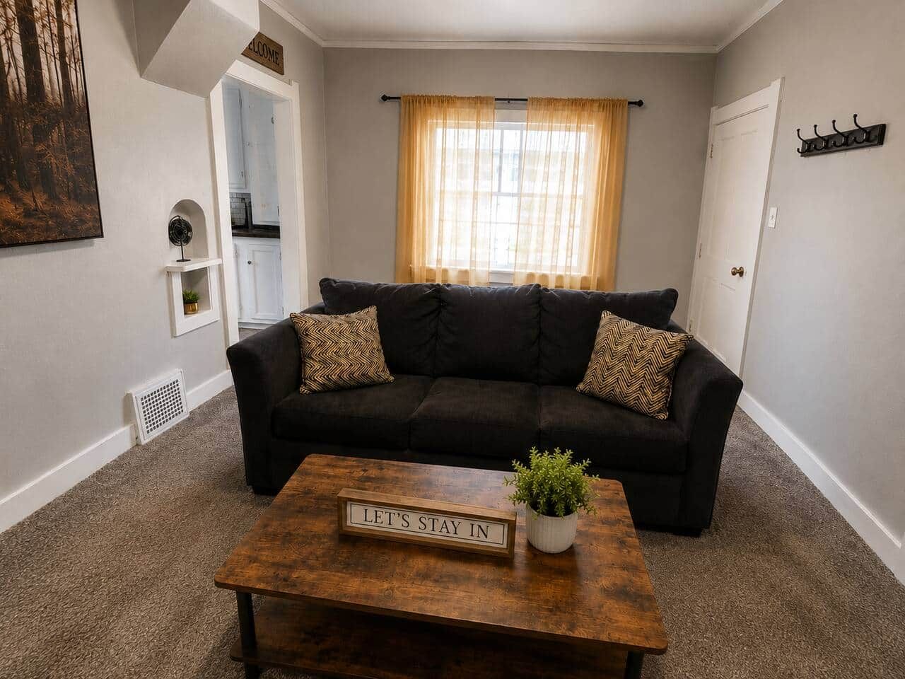 Ein Wohnzimmer mit einem dunkelgrauen Sofa, einem Couchtisch mit einem Schild und einem Fenster mit Vorhängen.