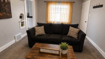 Una sala de estar con un sofá de color oscuro, una mesa de centro con un letrero y una ventana con cortinas.