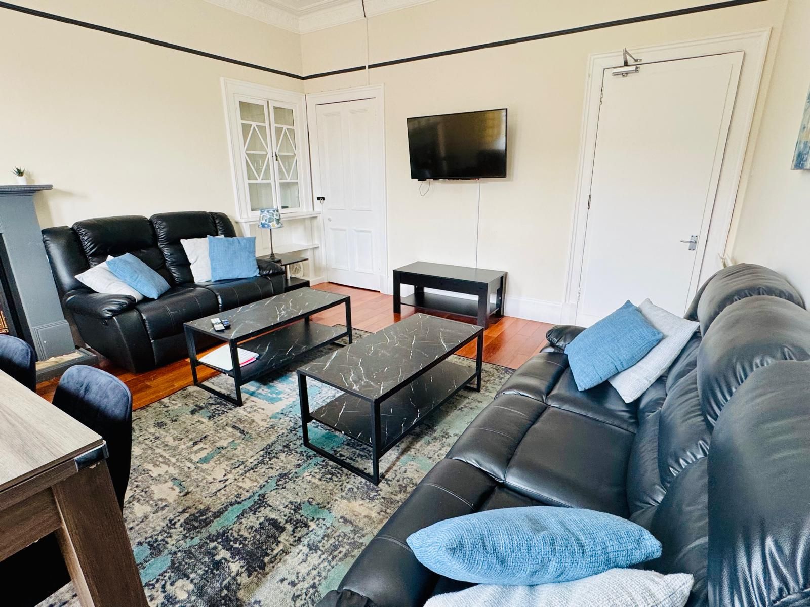 Ein Wohnzimmer mit einem schwarzen Ledersofa, einem Couchtisch, einem an der Wand montierten Fernseher und einer Tür mit Glasfenster.