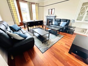 A living room with a sofa, coffee table, fireplace, and a dining area with chairs. - Spacious 4-Bed Duplex with Pool Table, Near GLA Airport (Renfrewshire)