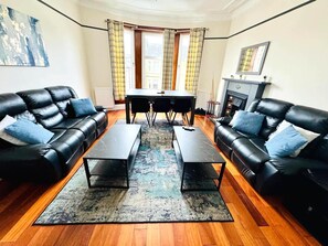 Ein Wohnzimmer mit zwei schwarzen Ledersofas, einem Couchtisch, einem Esstisch mit Stühlen, einem Kamin und einem großen Fenster mit Vorhängen.