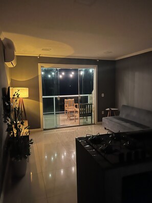 A modern living room with a sofa, a coffee table, and a dining table. There is a sliding glass door leading to an outdoor area with chairs.