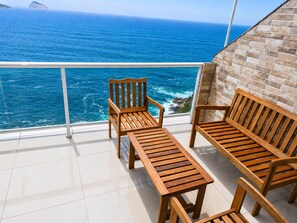 A balcony with two wooden chairs and a table, overlooking a body of water with a rocky coastline.