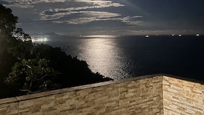 Vista nocturna de la costa con una luna brillante, mar tranquilo y luces de la ciudad a lo lejos.