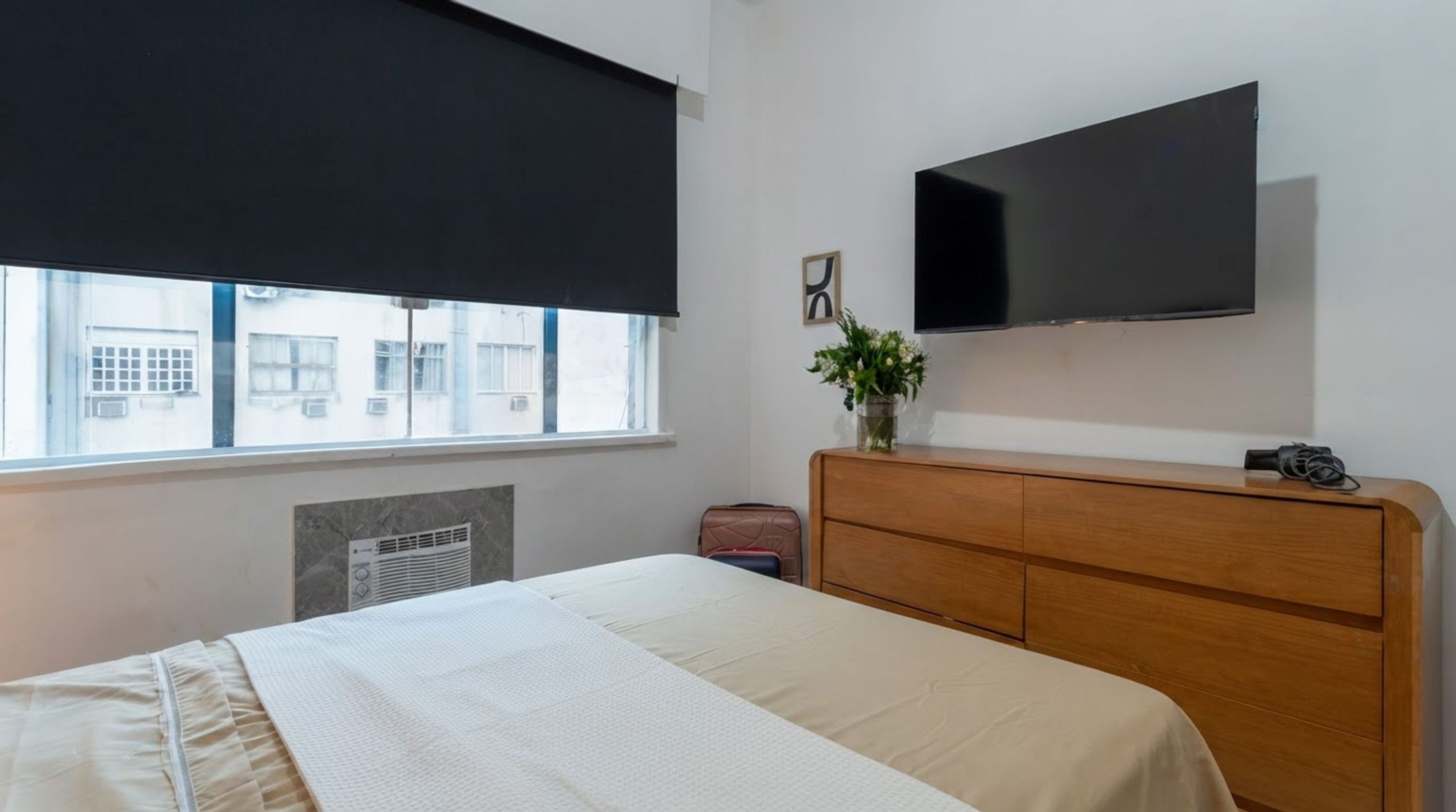 Een hotelkamer met een bed, een houten dressoir, een flatscreen televisie, een raam met uitzicht op gebouwen en een radiator.