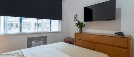 A hotel room with a bed, a wooden dresser, a flat-screen TV, a window with a view of buildings, and a radiator.