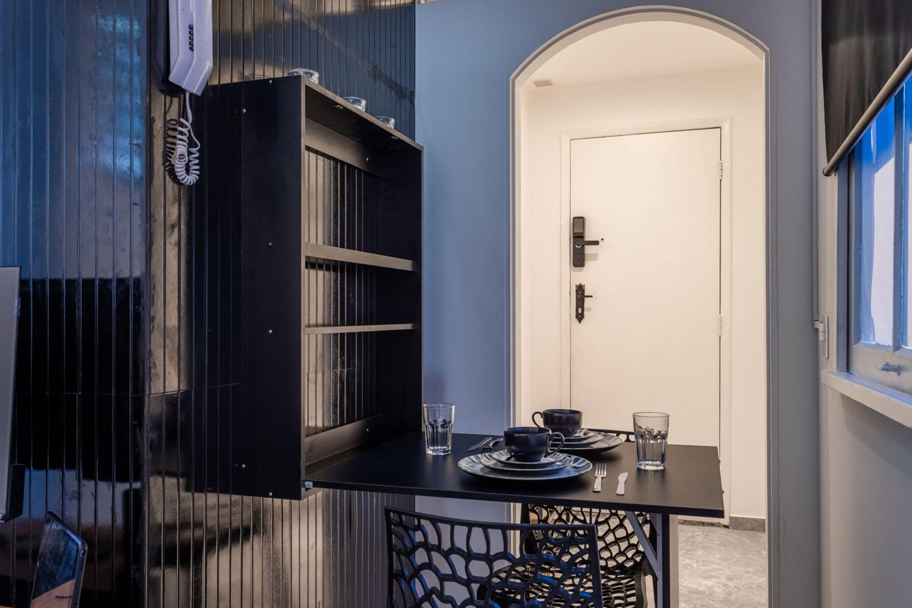 A compact room with a dining table set for two, a door with a modern lock, and a window with blinds.