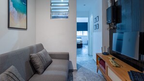 Una sala de estar moderna con un sofá gris, un televisor de pantalla plana montado en un panel de madera, y una pequeña mesa con un teclado y un ratón.