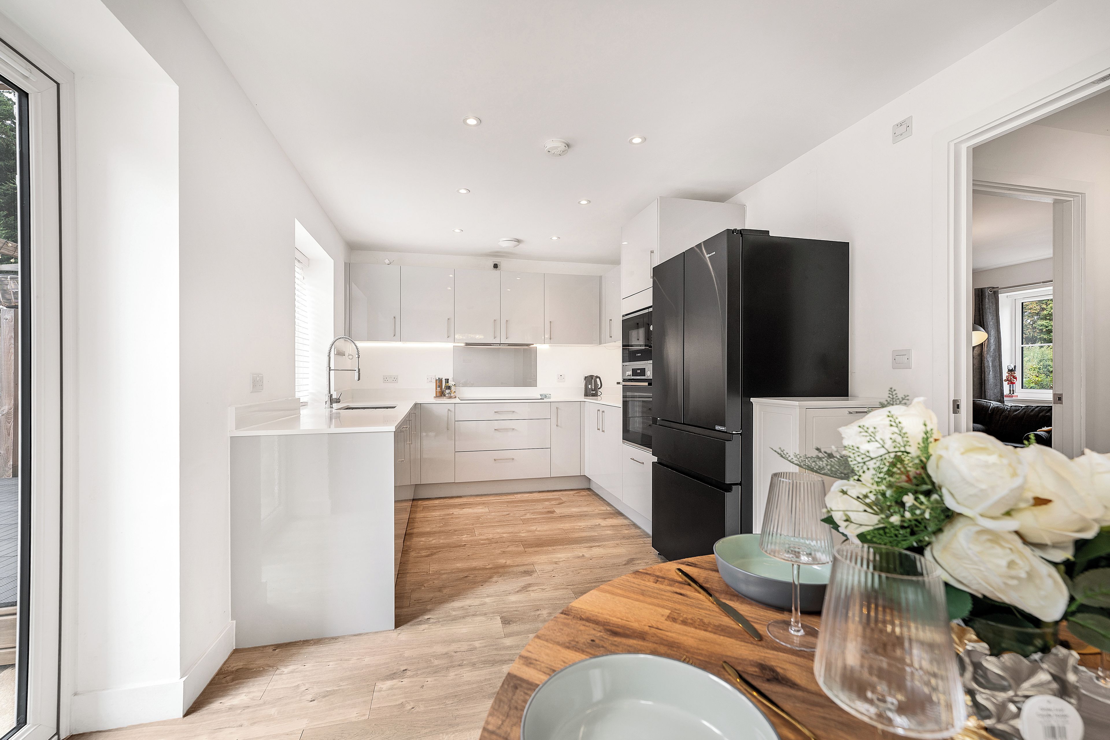 A modern kitchen with white cabinetry, a black refrigerator, and a wooden dining table with a vase of flowers.