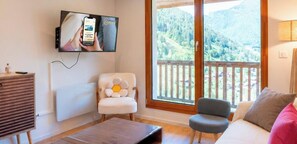 Ein Wohnzimmer mit an der Wand montiertem Fernseher, einer Couch, einem Sessel und Blick auf eine gebirgige Landschaft durch einen Balkon.