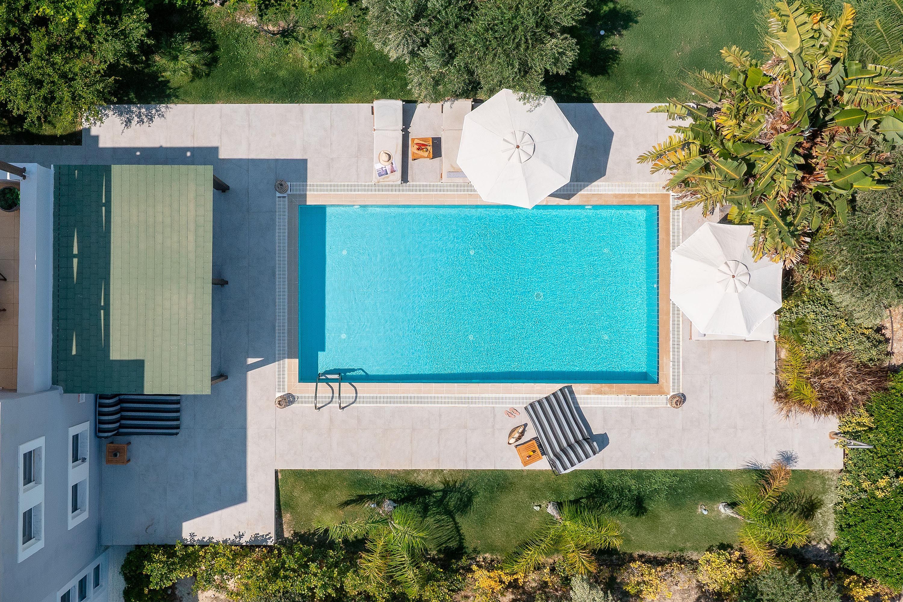 An aerial view of a rectangular swimming pool with a surrounding deck, a covered seating area, and a small grassy area.