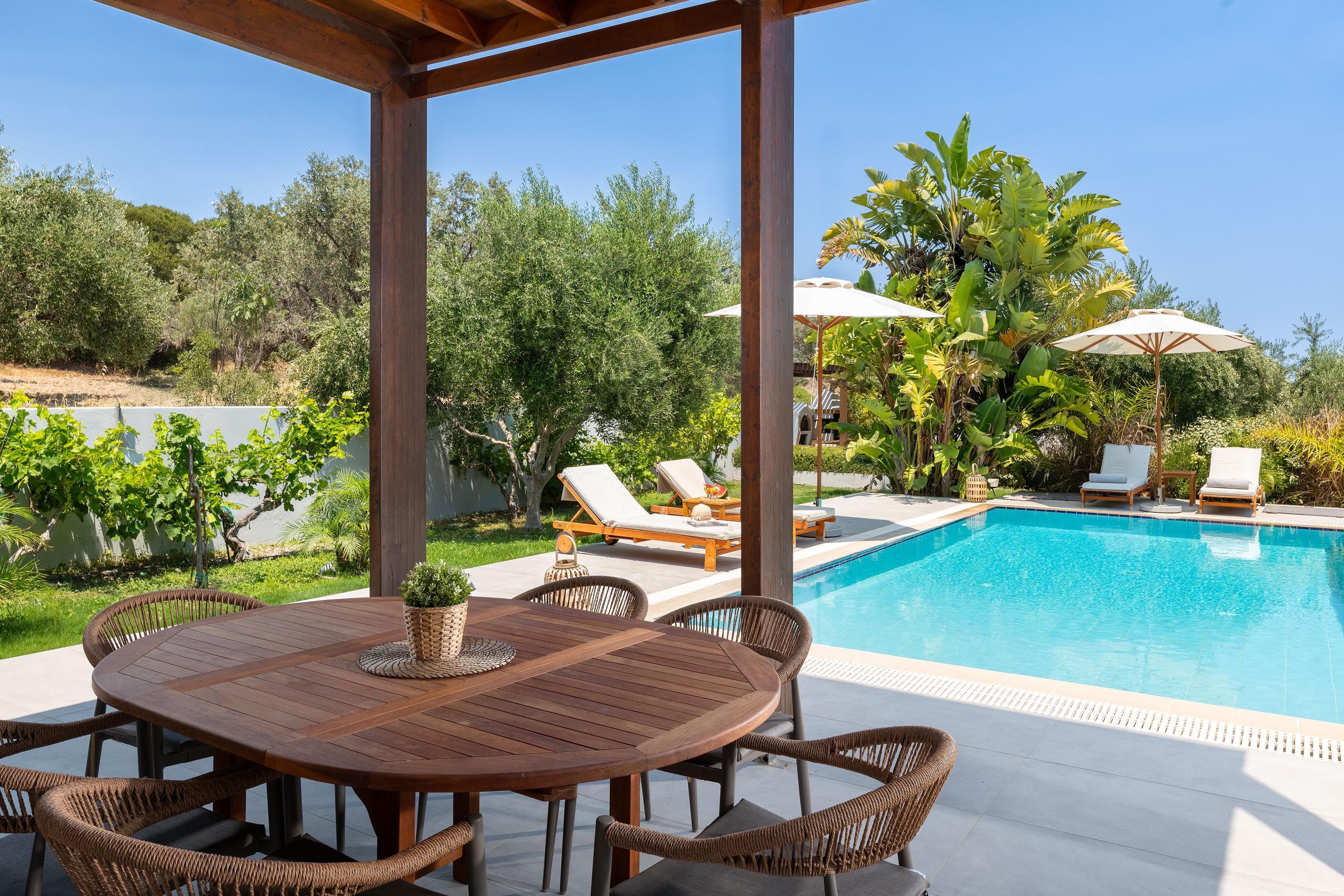Un espace aménagé au bord de la piscine, avec une table en bois et des chaises en osier, entouré de transats et de parasols.