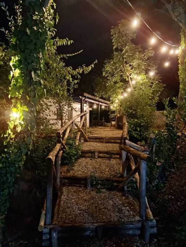 A wooden staircase leading to a building, surrounded by greenery and illuminated by string lights.