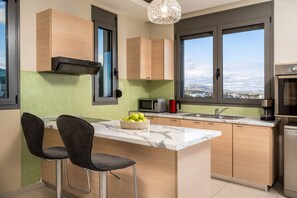 A modern kitchen with a central island, two bar stools, a microwave, a coffee maker, and a view of the city through large windows. - Villa Aqua in Lachania (Lachania)
