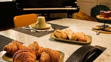 A dining table with croissants and a cake, a piano in the background, and a window with a view.