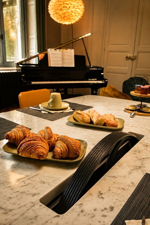 A dining table with croissants and a cake, a piano in the background, and a window with a view.