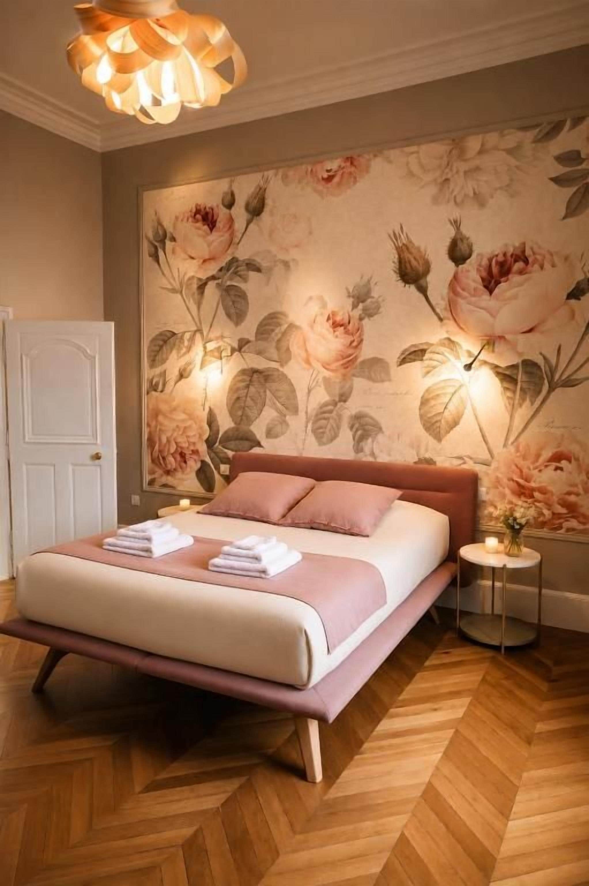 Un dormitorio con cama, mesita de noche, lámpara de pared y papel tapiz floral.