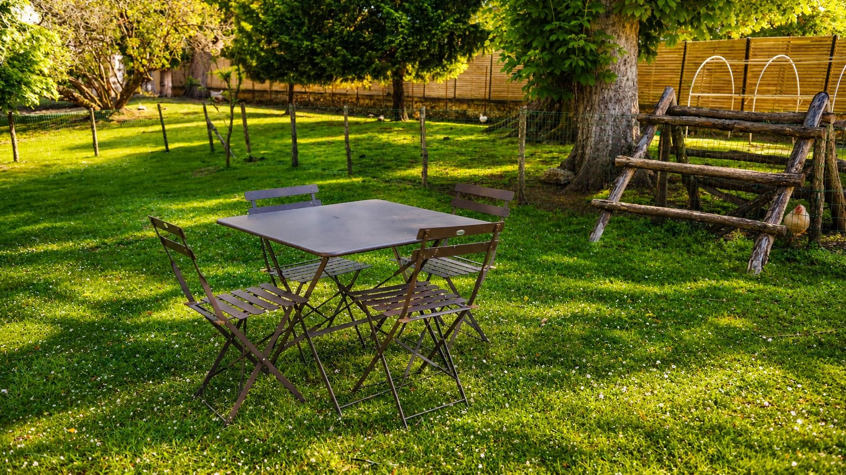 An outdoor seating area with a metal table and chairs, surrounded by a grassy lawn and trees.