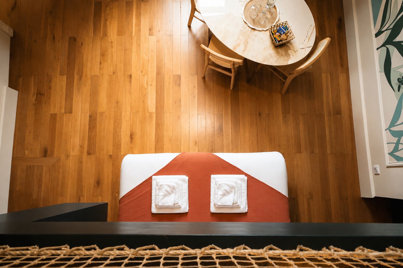 Une pièce avec un plancher en bois, une table ronde entourée de chaises, un lit recouvert d’une couvre-lit rouge et blanche, et un tapis tissé sur le sol.