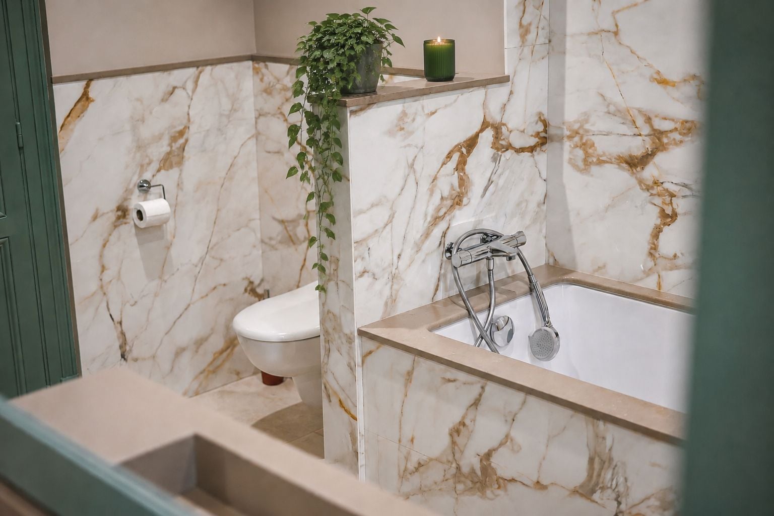 A bathroom with a marble wall, a toilet, a bathtub with a showerhead, and a green plant on a shelf.
