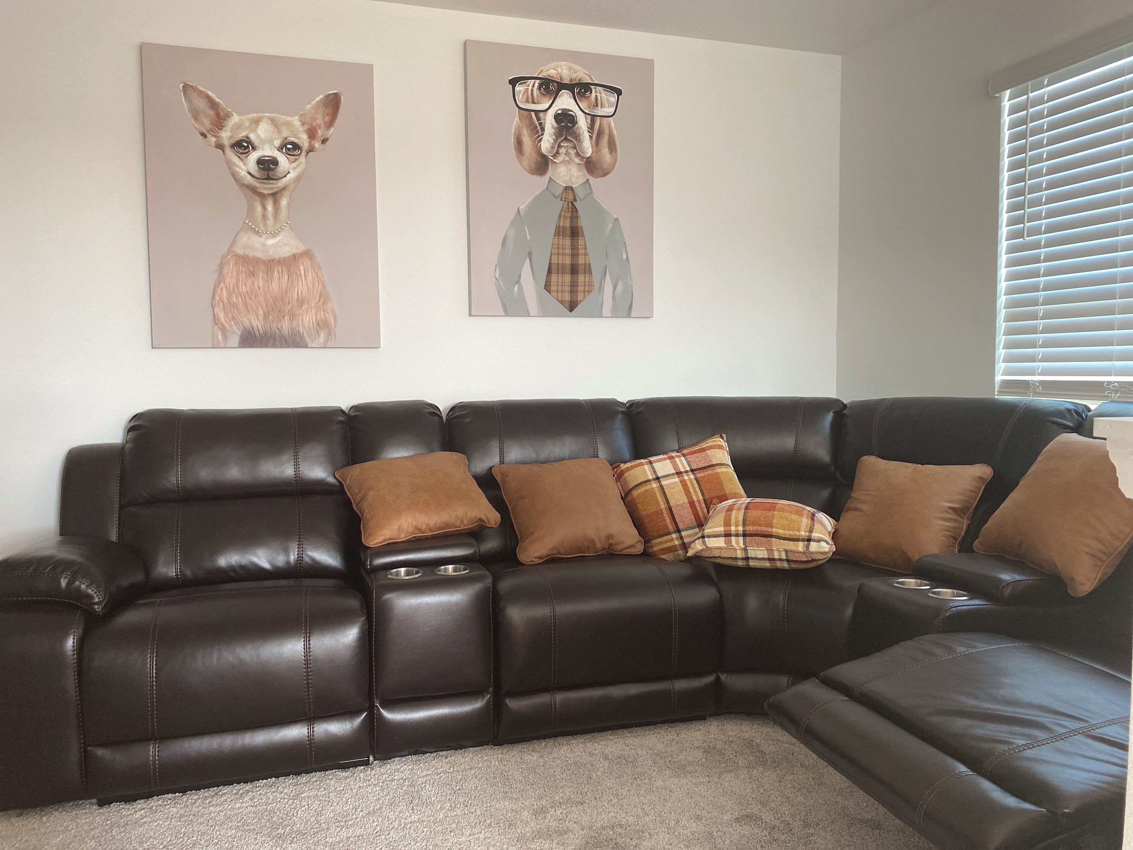 A dark brown leather sectional sofa with multiple cushions, a carpeted floor, and two wall-mounted dog portraits.