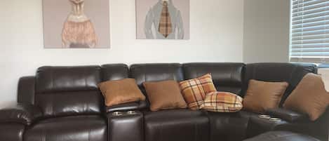 A dark brown leather sectional sofa with multiple cushions, a carpeted floor, and two wall-mounted dog portraits.