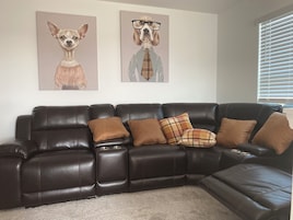 A dark brown leather sectional sofa with multiple cushions, a carpeted floor, and two wall-mounted dog portraits.