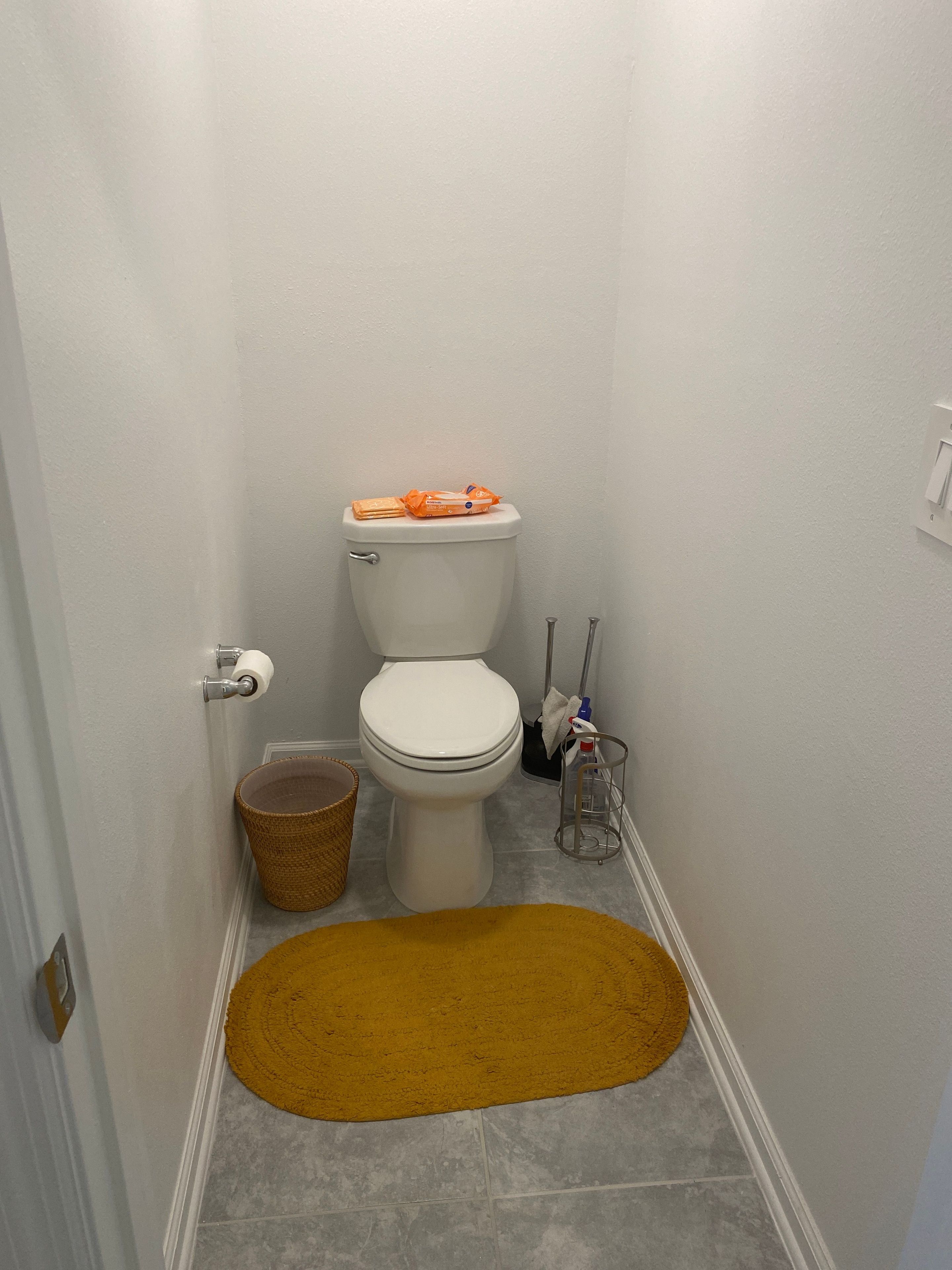 Un baño pequeño con inodoro, una cesta de mimbre, un tapete amarillo y artículos de limpieza.