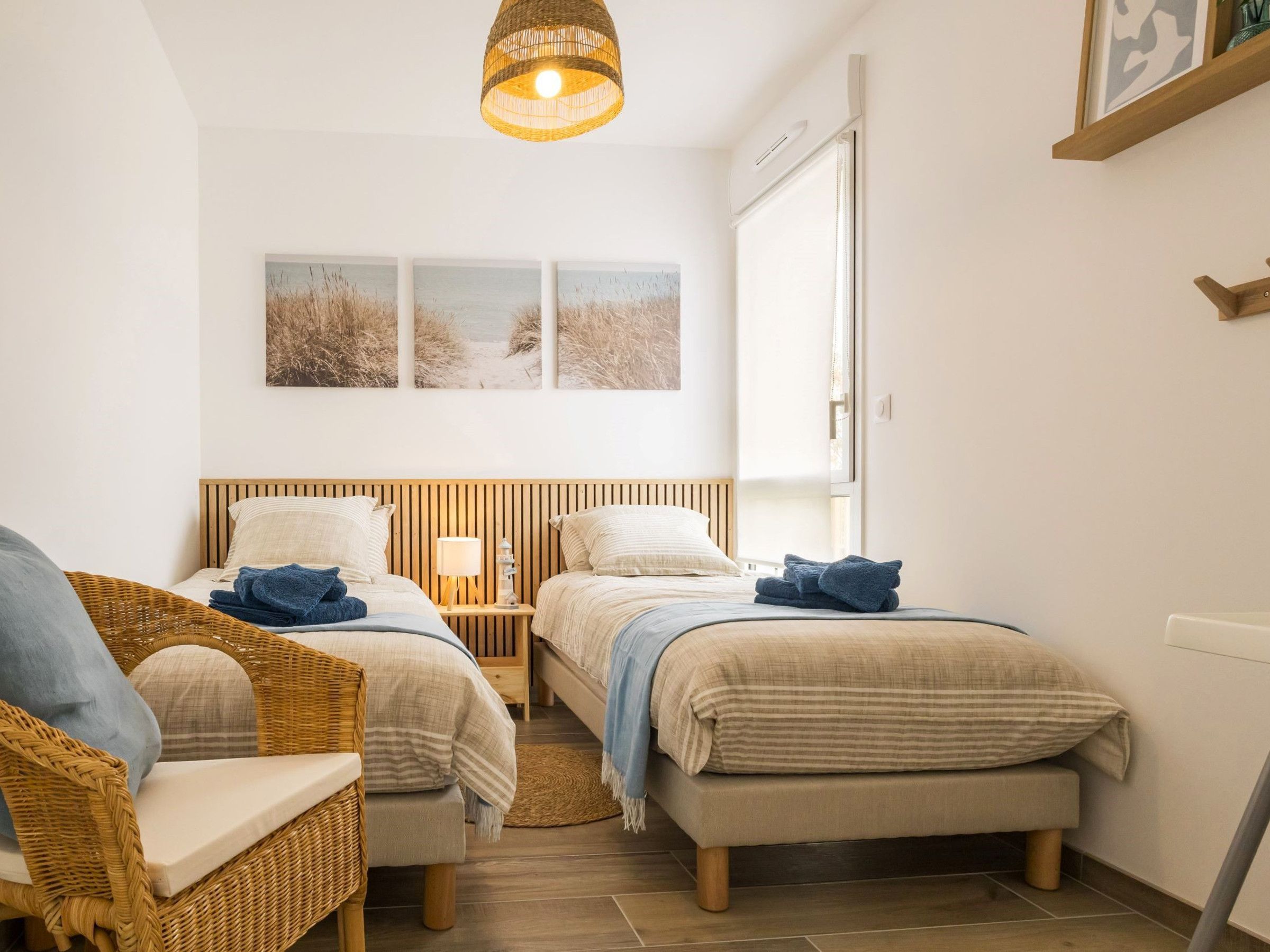 Une chambre avec deux lits, une chaise en osier, une table de chevet avec une lampe et une fenêtre donnant sur un beau paysage.