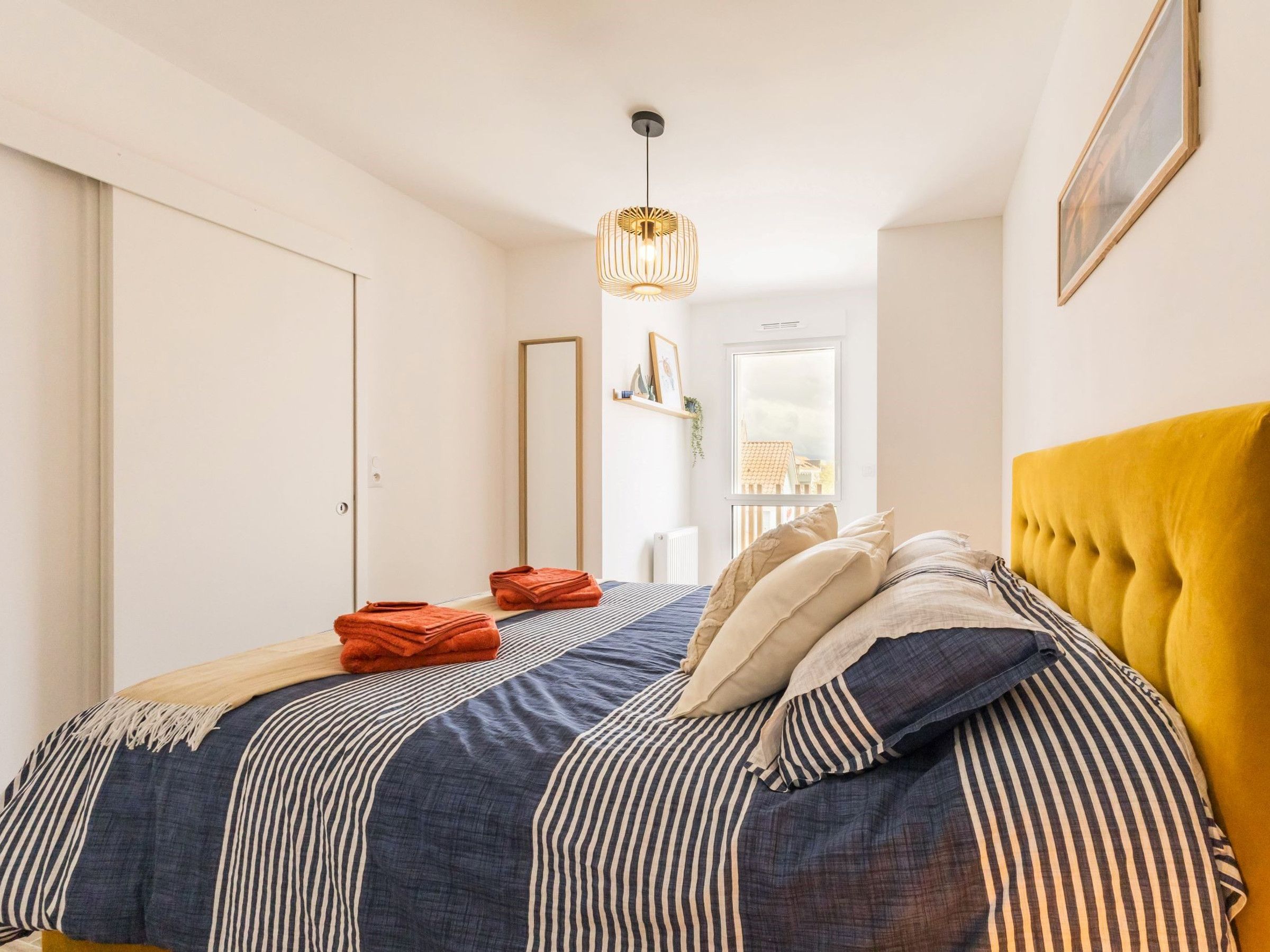 A bedroom with a bed, a mirror, a window, and a hanging light fixture.